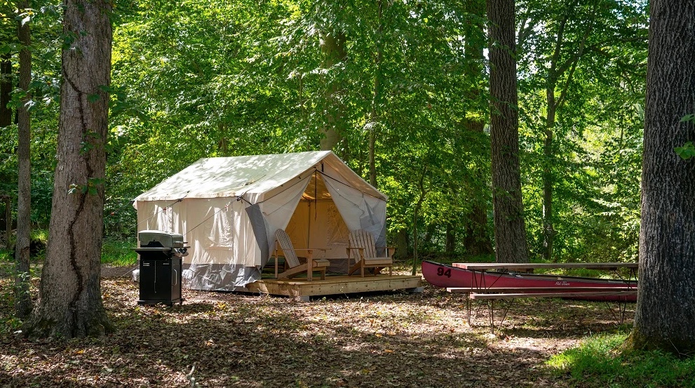 Tent in forest with deck chairs, grill, canoe, and picnic table under trees.