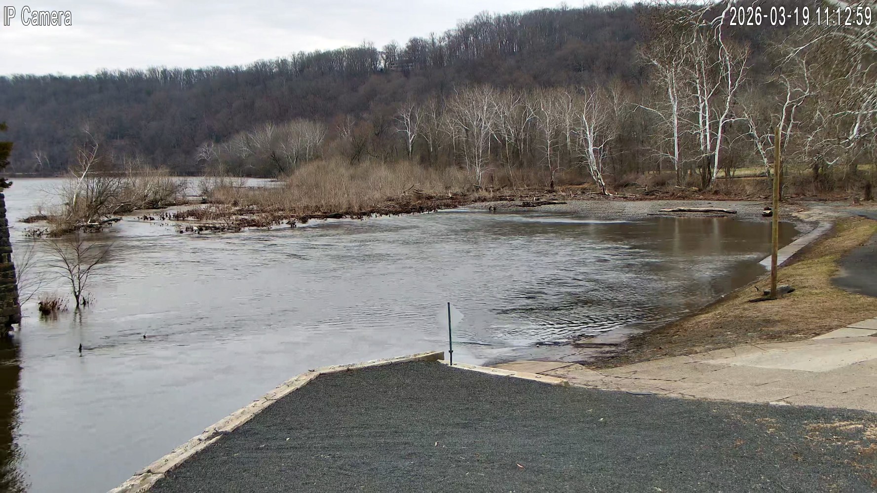 Flooded riverbank with trees and a paved path on a cloudy day.